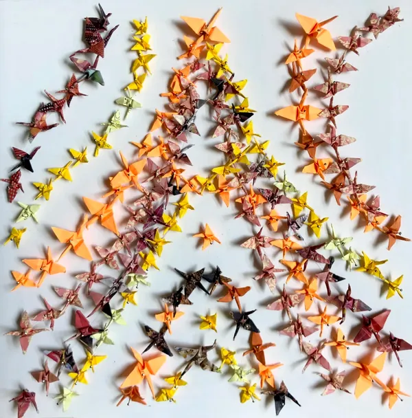 small paper cranes in shades of yellow and orange arranged in lines following the shape of an A, pinned to a white background