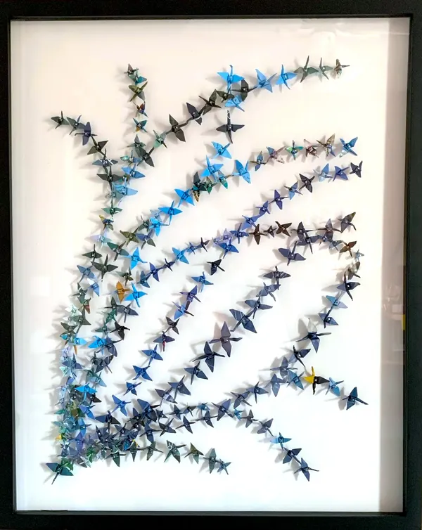 small paper cranes in shades of blue and teal pinned to white background, arranged in wavy lines starting from the bottom left corner